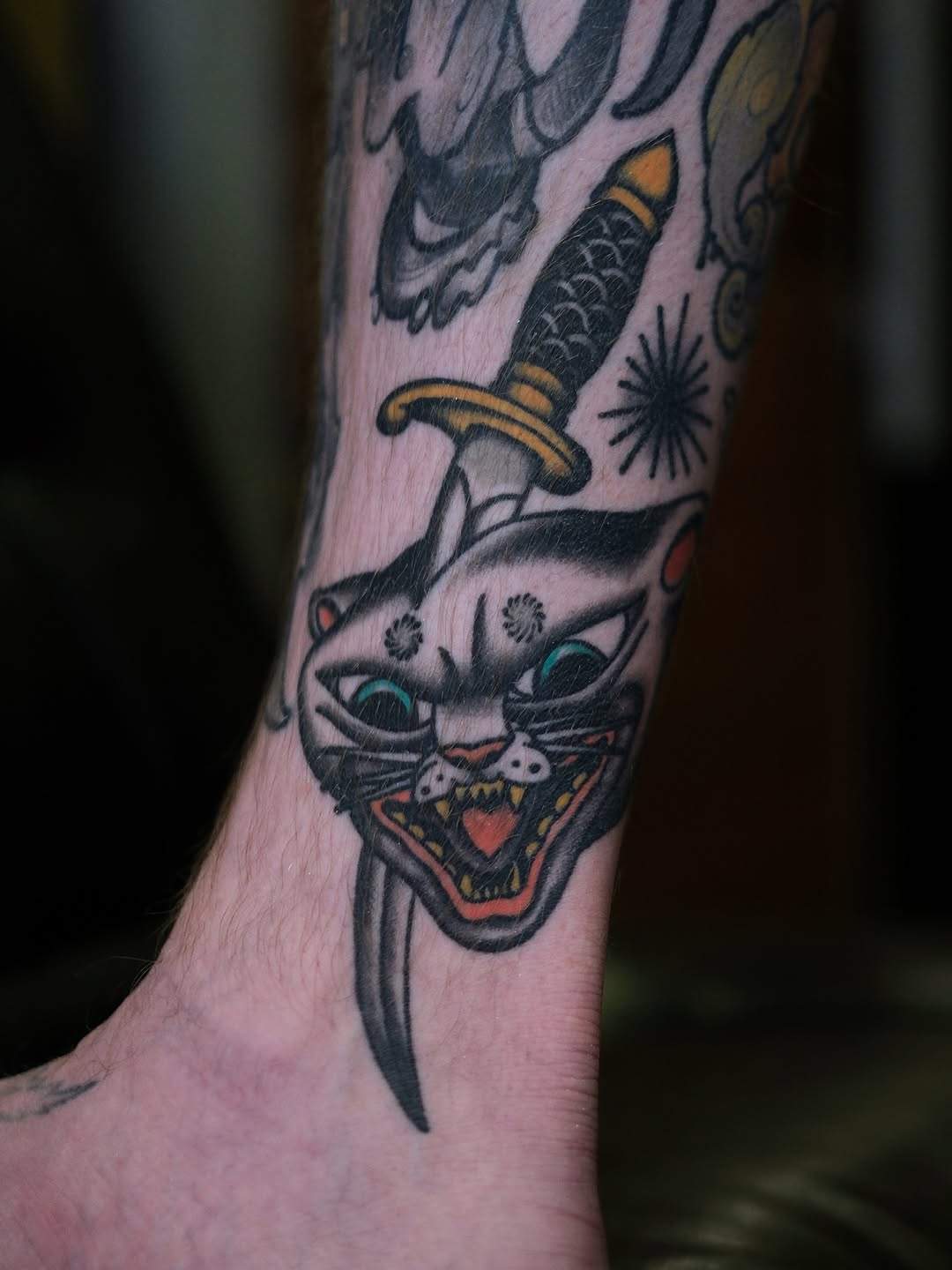 Cat head with glowing blue eyes and open mouth holding a dagger with gold handle, tattooed on the ankle in black, gold, and red ink.