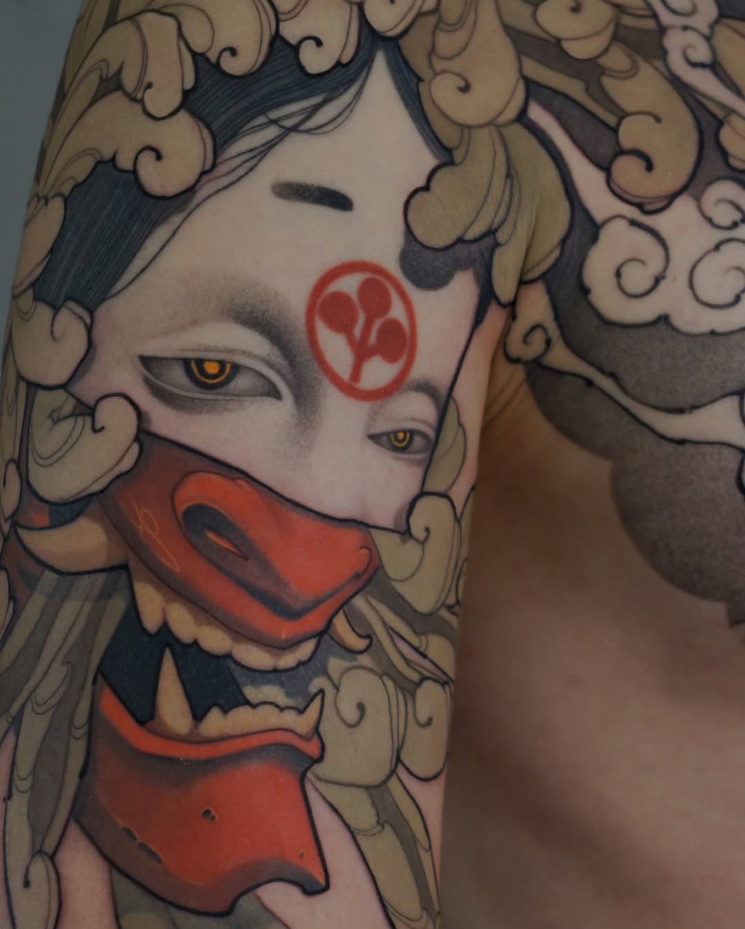Tattoo on upper arm featuring a stylized face with golden eyes, red facial markings, ornate curled hair details in tan and black, and red mouth covering.