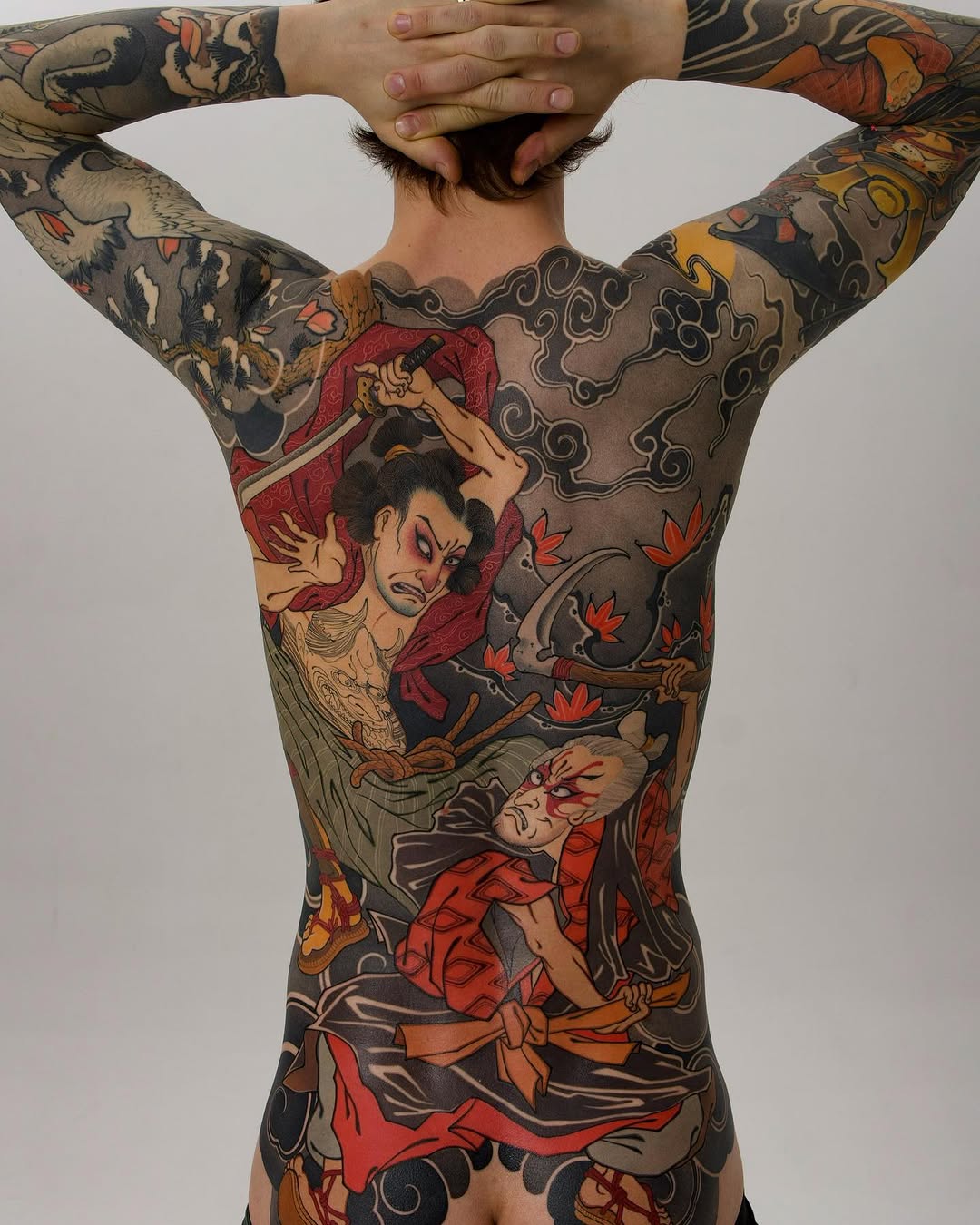 Rear view of a person's back and arms covered in colorful tattoos featuring mythological and fantastical characters in red, black, tan, and orange hues.