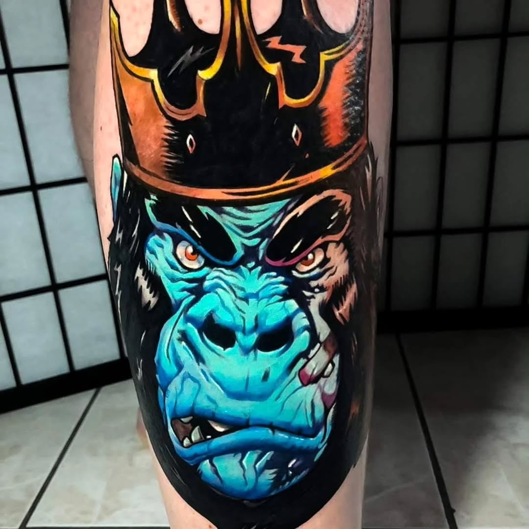Colorful demon face tattoo on leg featuring bright blue skin, orange flames, glowing eyes, and bared teeth against black background.