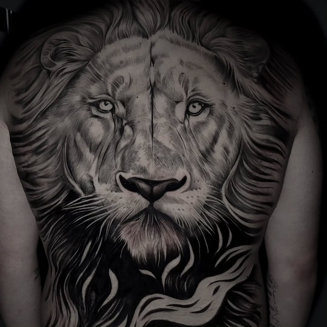 Large lion head tattoo on chest and upper back with detailed mane, frontal gaze, and flowing hair elements rendered in black and grey ink.