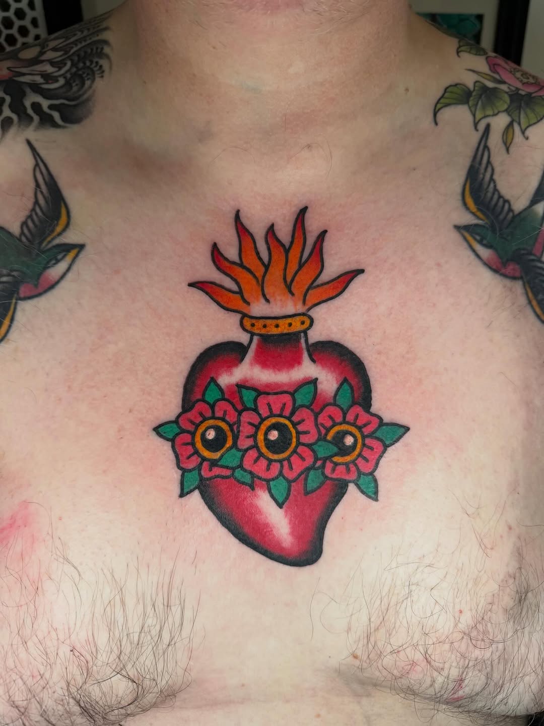 Sacred heart tattoo with flame crown, three red flowers with yellow centers, and green leaves on the chest.