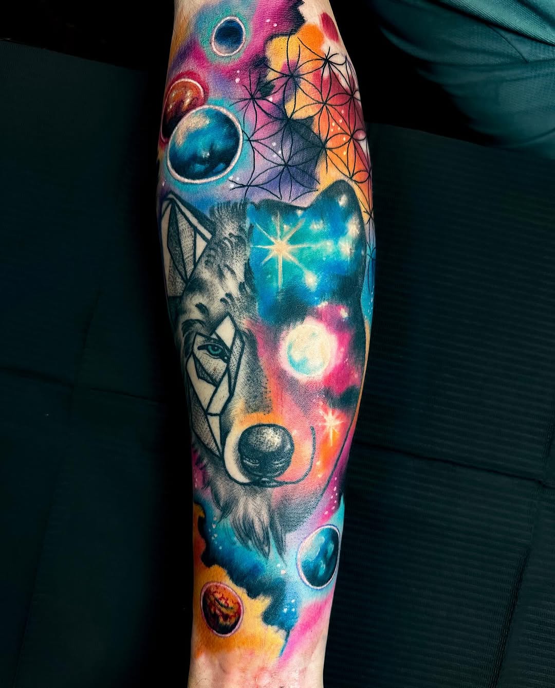 Colorful forearm tattoo featuring a wolf's head with geometric patterns, celestial bodies, planets, and star symbols in vibrant blues, pinks, oranges, and yellows.