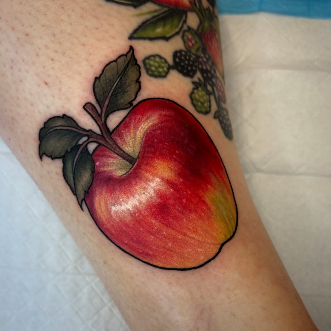 Colorful apple tattoo with green leaves on the upper arm, featuring red, orange, and yellow shading with black outlines.