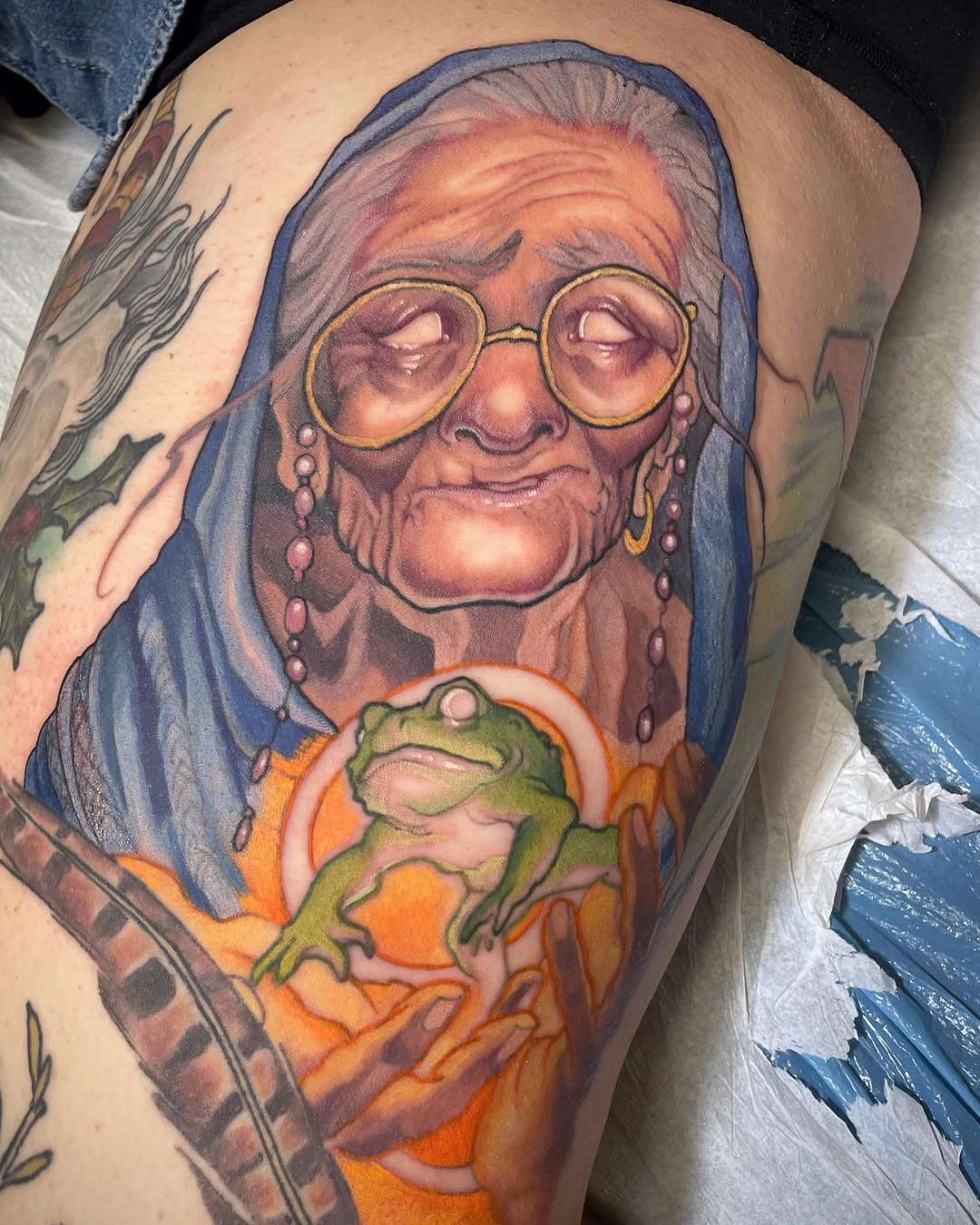 Colorful shoulder tattoo depicting an elderly woman wearing glasses and blue headscarf holding a green frog, with orange and yellow background tones.