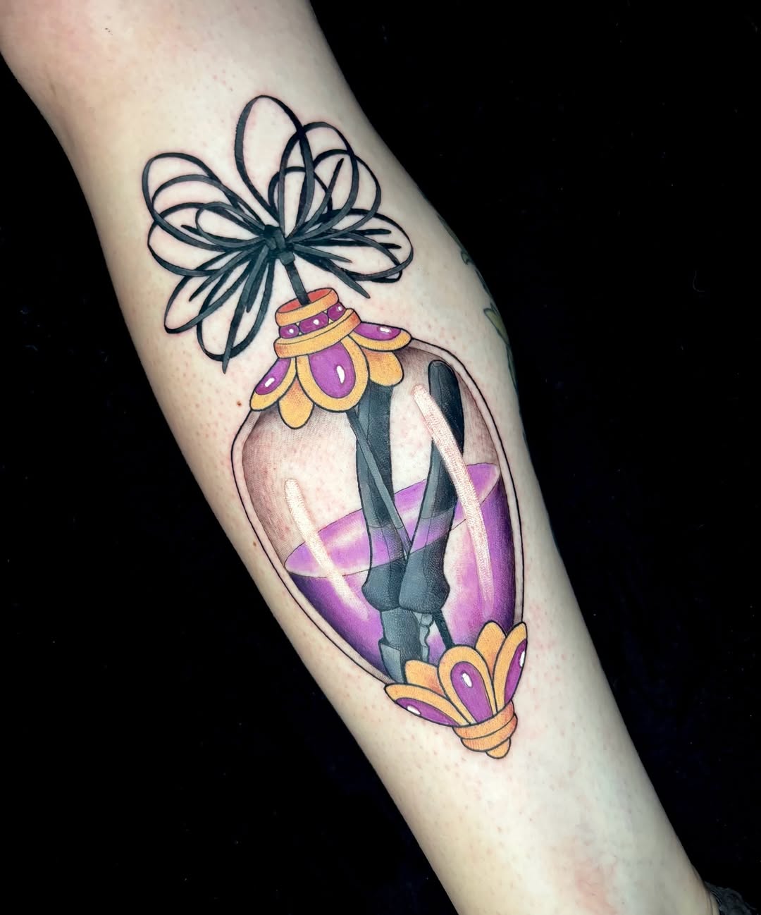 Colorful tattoo on an arm featuring a decorative perfume bottle with purple and gold accents, topped with a dark blue bow.