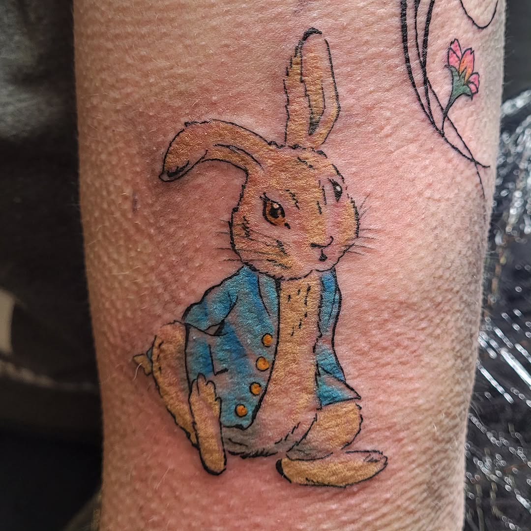 Colored tattoo of a rabbit wearing a blue jacket with orange buttons on the forearm, with small flowers and vines in the background.