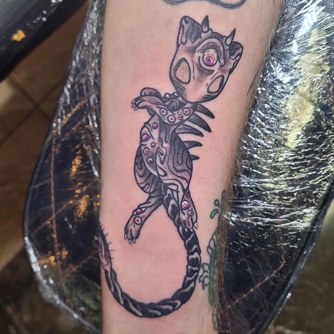 Fantastical creature tattoo with spotted head, spiky mane, decorative body markings, and curled tail on an upper arm in black ink with purple accents.