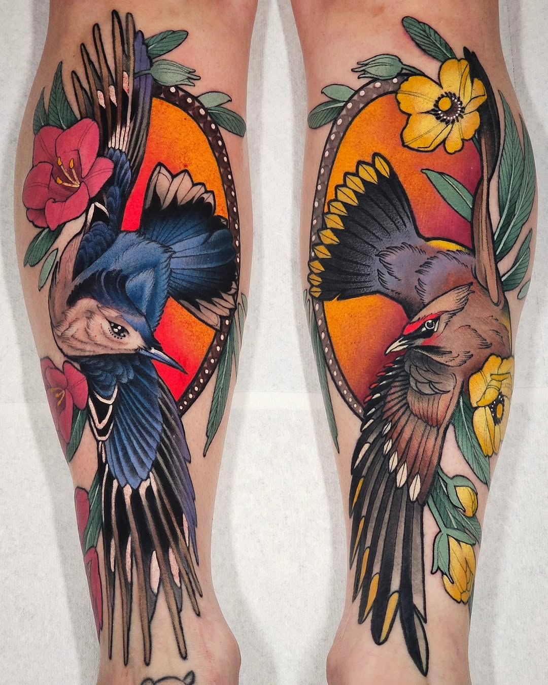 Matching pair of colorful bird tattoos on both calves featuring blue and brown birds with red and orange circular backgrounds, surrounded by pink and yellow flowers with green leaves.