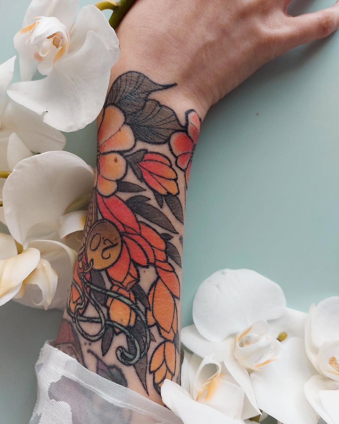 Colorful floral tattoo on the forearm featuring red, orange, and yellow flowers with black linework and leaves, displayed against white orchids.