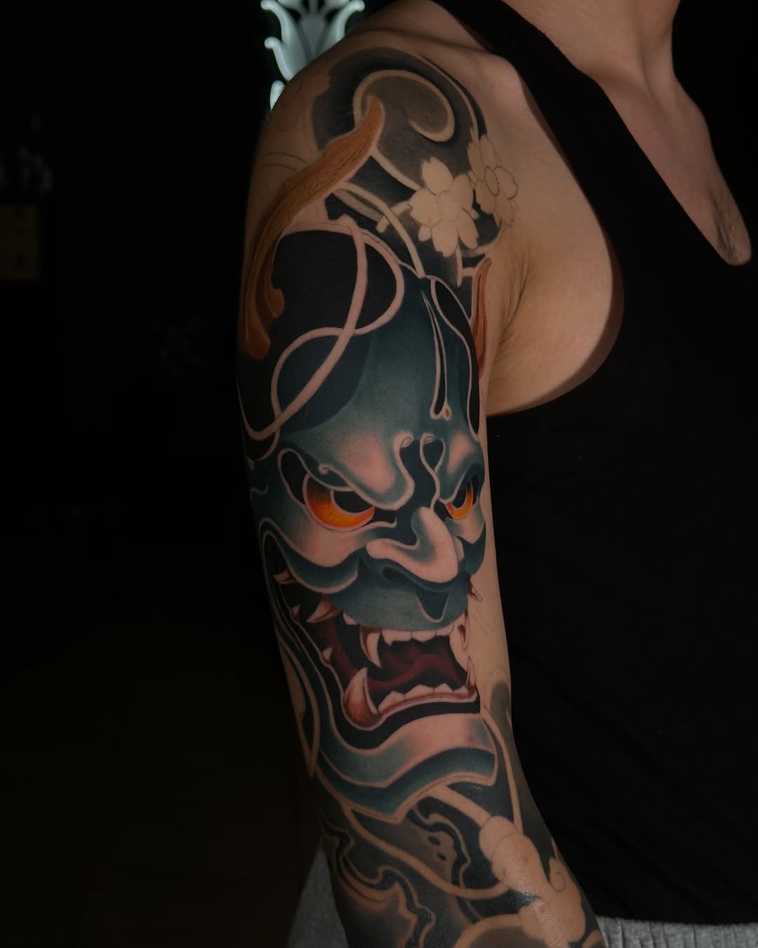 Demon mask tattoo on upper arm and shoulder featuring glowing orange eyes, ornate horns, and floral vine elements in black, orange, and skin tone colors.