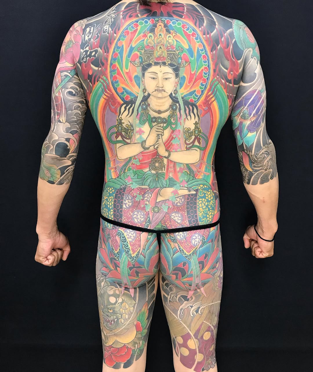 Back view of a full-body tattoo featuring a deity figure with ornate headdress in the center, surrounded by colorful religious imagery, floral patterns, and decorative elements in red, green, gold, and blue.