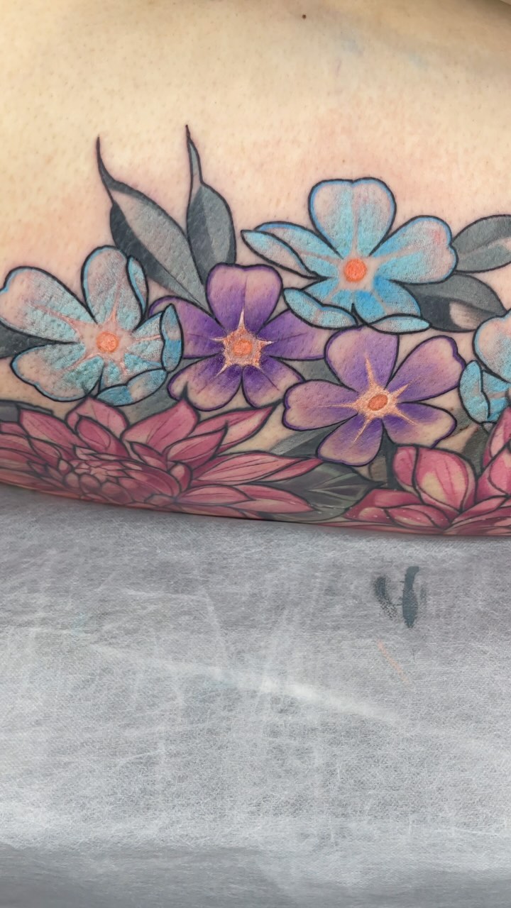 Colorful floral tattoo on forearm featuring blue, purple, and pink flowers with orange centers and green leaves arranged horizontally.