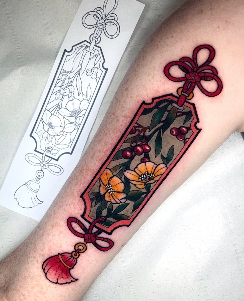 Colorful tattoo on forearm featuring a red-bordered ornamental frame with orange flowers, red berries, green leaves, and decorative red bows at top and bottom.