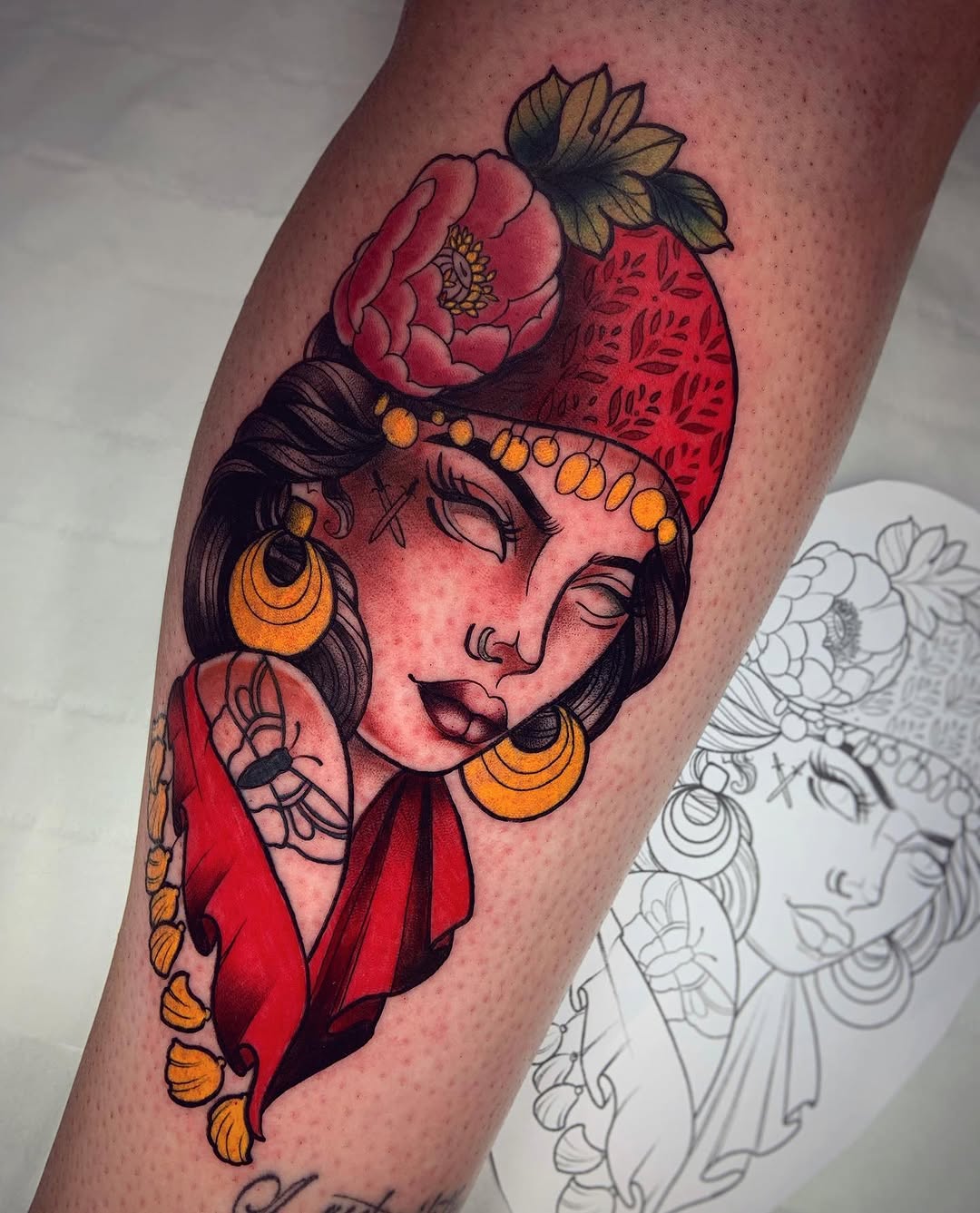 Colorful tattoo on a calf featuring a woman's face with gold hoop earrings, red clothing, and a red peony with yellow leaves positioned above her head.