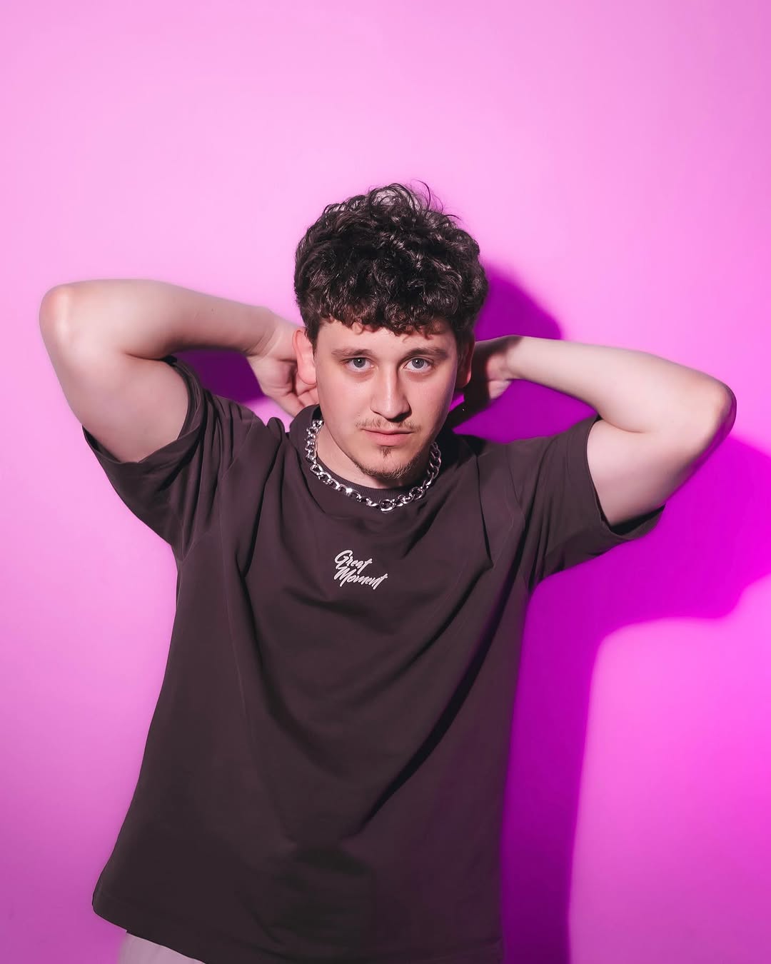 A young man wearing a dark t-shirt and chain necklace displays a small tattoo on his upper chest against a bright pink background.