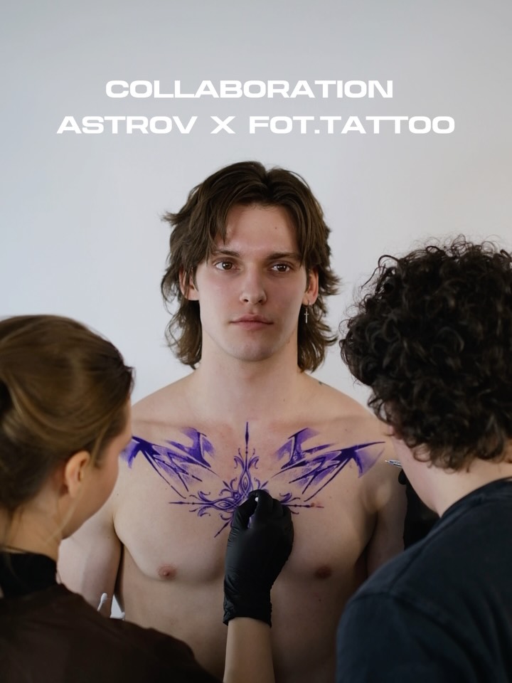 Tattoo artist applying purple and black chest piece featuring geometric arrows and ornamental details to a shirtless male client.