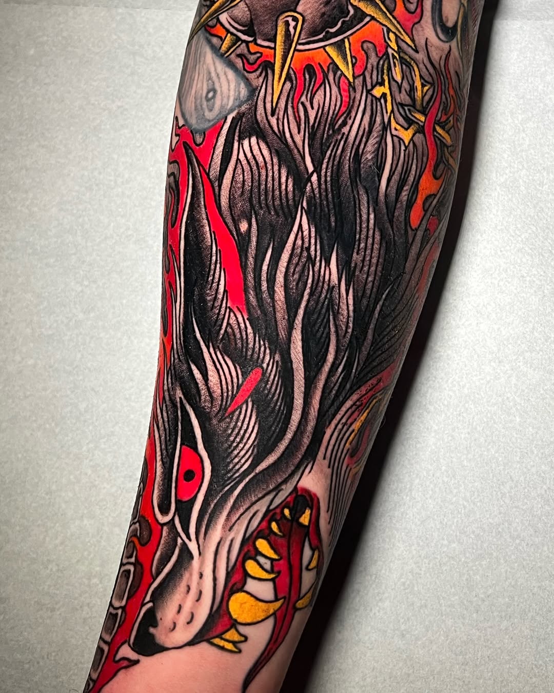 Colorful tattoo on a forearm featuring a bearded figure with flowing hair surrounded by red flames, yellow accents, and ornamental details.
