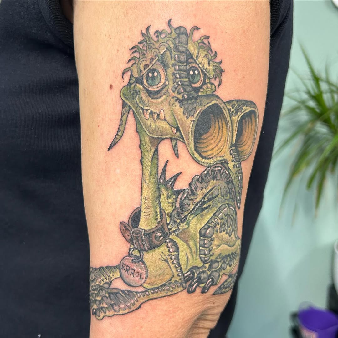 Colorful tattoo on upper arm featuring a grotesque creature with wild hair, large eyes, mechanical elements, and a tag reading "ERROL" in yellow and black ink.