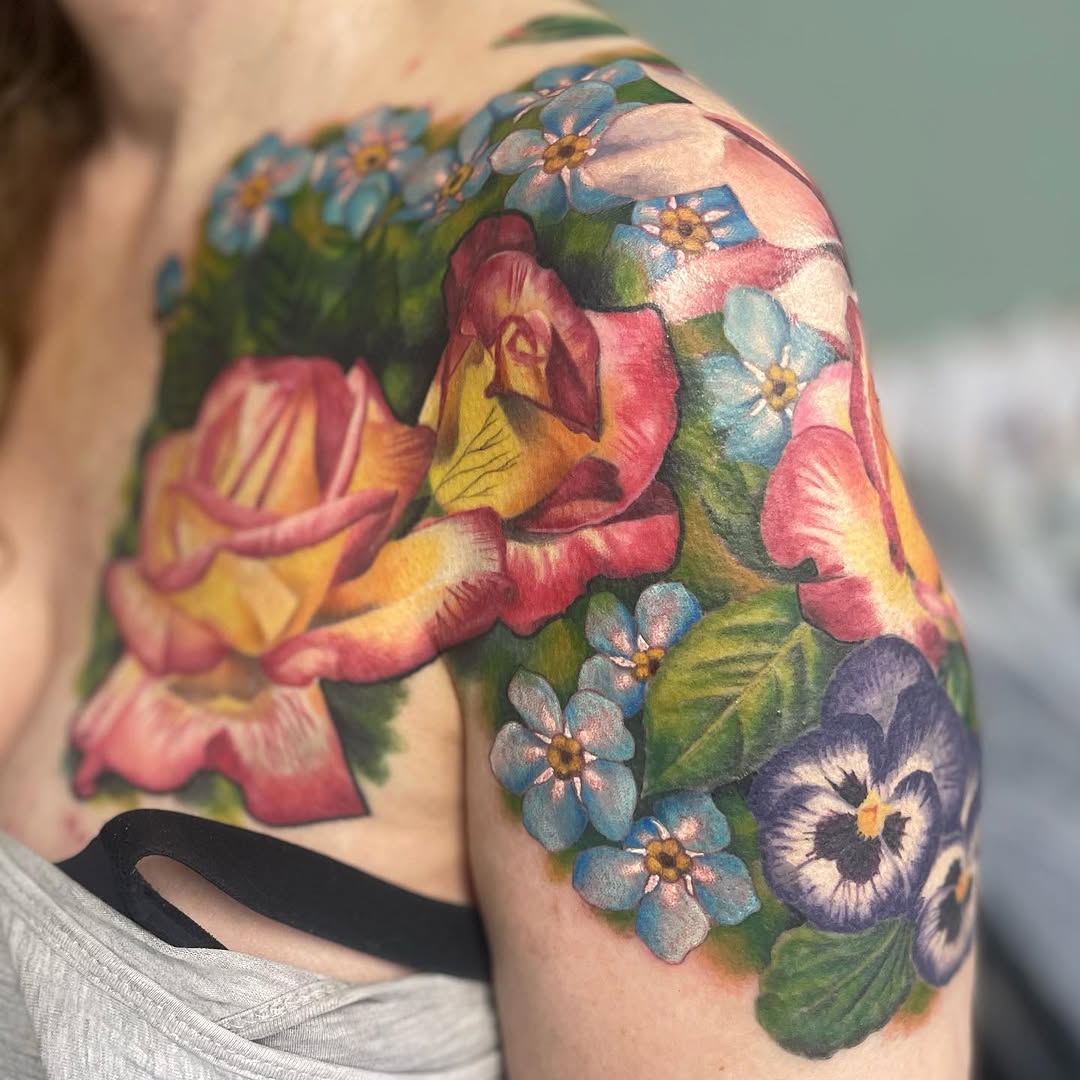 Colorful floral tattoo on upper arm featuring pink and yellow roses, blue forget-me-nots, and purple pansies with green leaves.