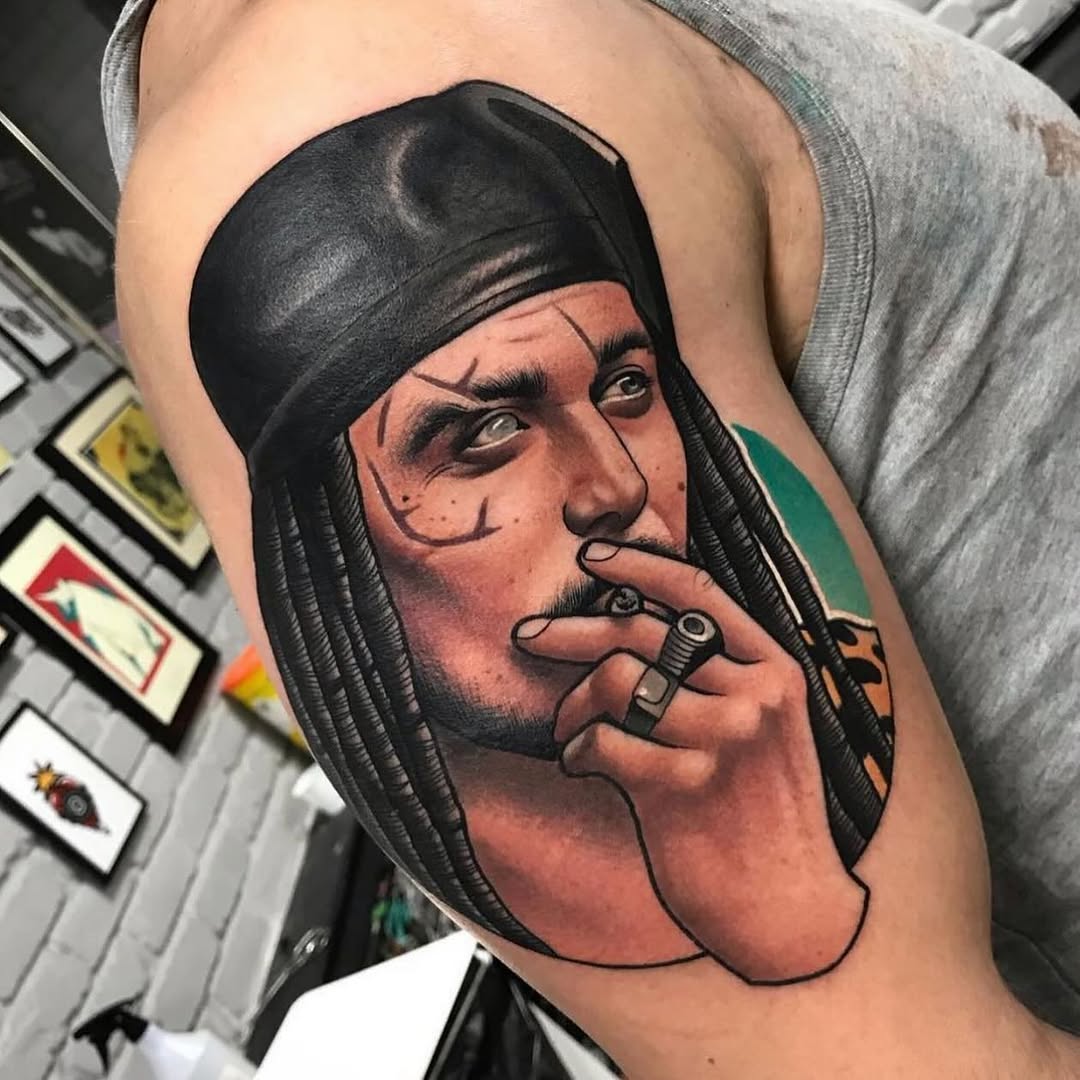 Colored portrait tattoo on upper arm depicting a man with long black hair and head wrap, smoking a cigarette, with green and orange accents.