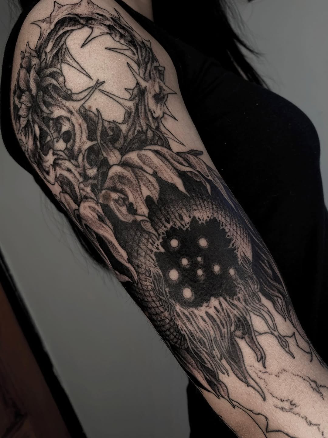 Detailed monochromatic arm tattoo featuring a bearded face with floral elements and an eye with dotted pupil, spanning from shoulder to forearm.