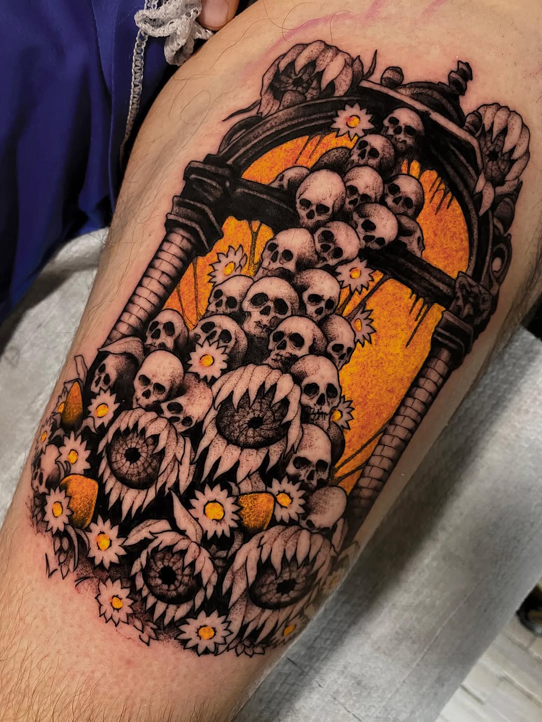 Colorful shoulder tattoo featuring skulls, eyes, sunflowers, and a scythe against an orange background with decorative foliage.