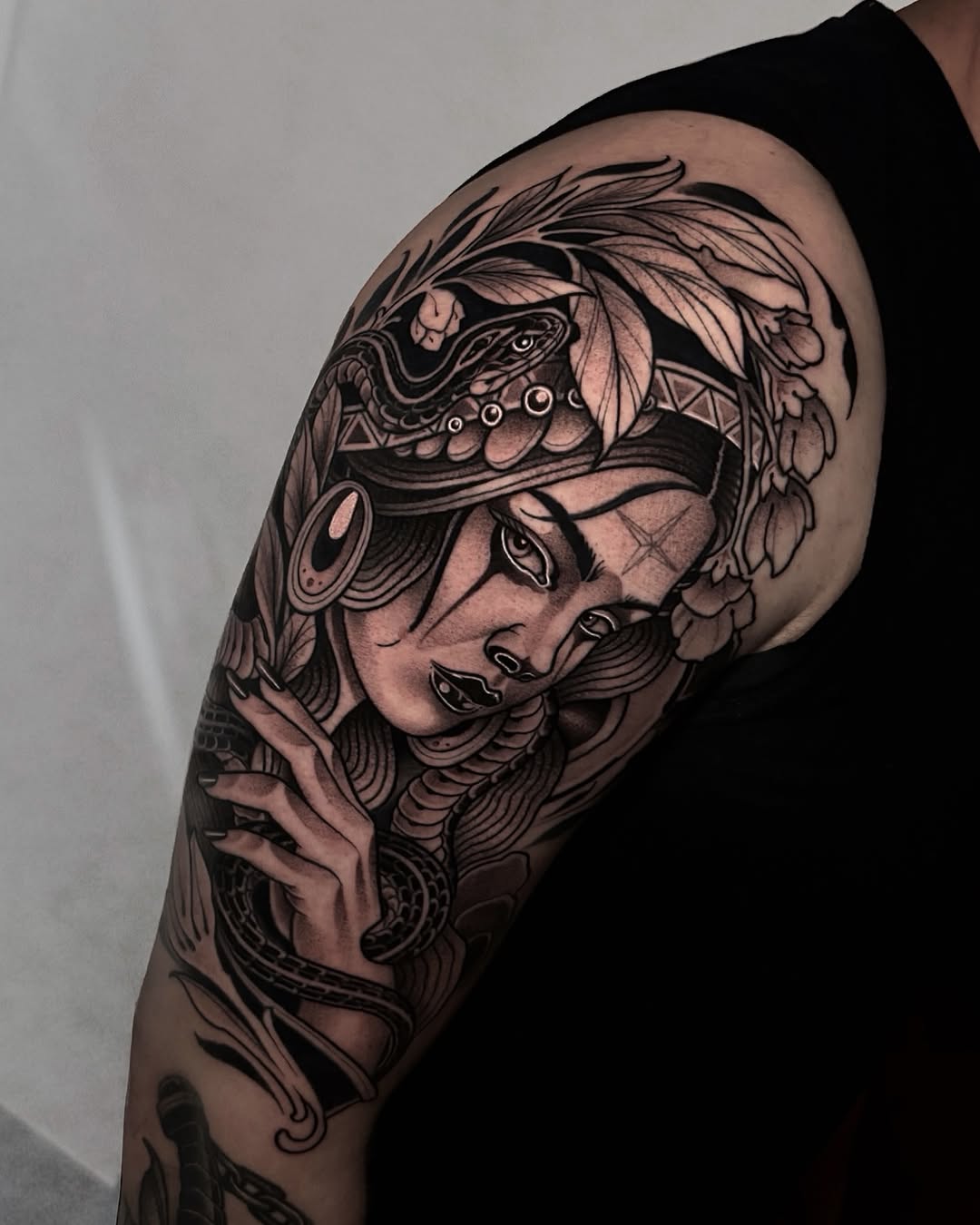 Detailed black ink portrait tattoo on upper arm featuring a face with ornate headdress, feathered elements, and decorative patterns covering the shoulder.