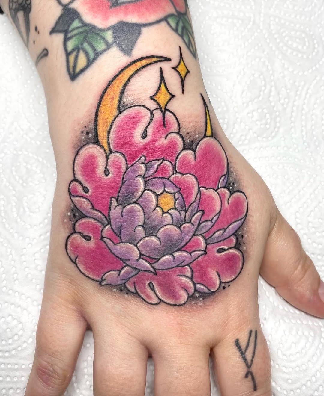 Colorful tattoo on a hand featuring a large pink and purple layered flower with a crescent moon and stars in gold and orange tones.