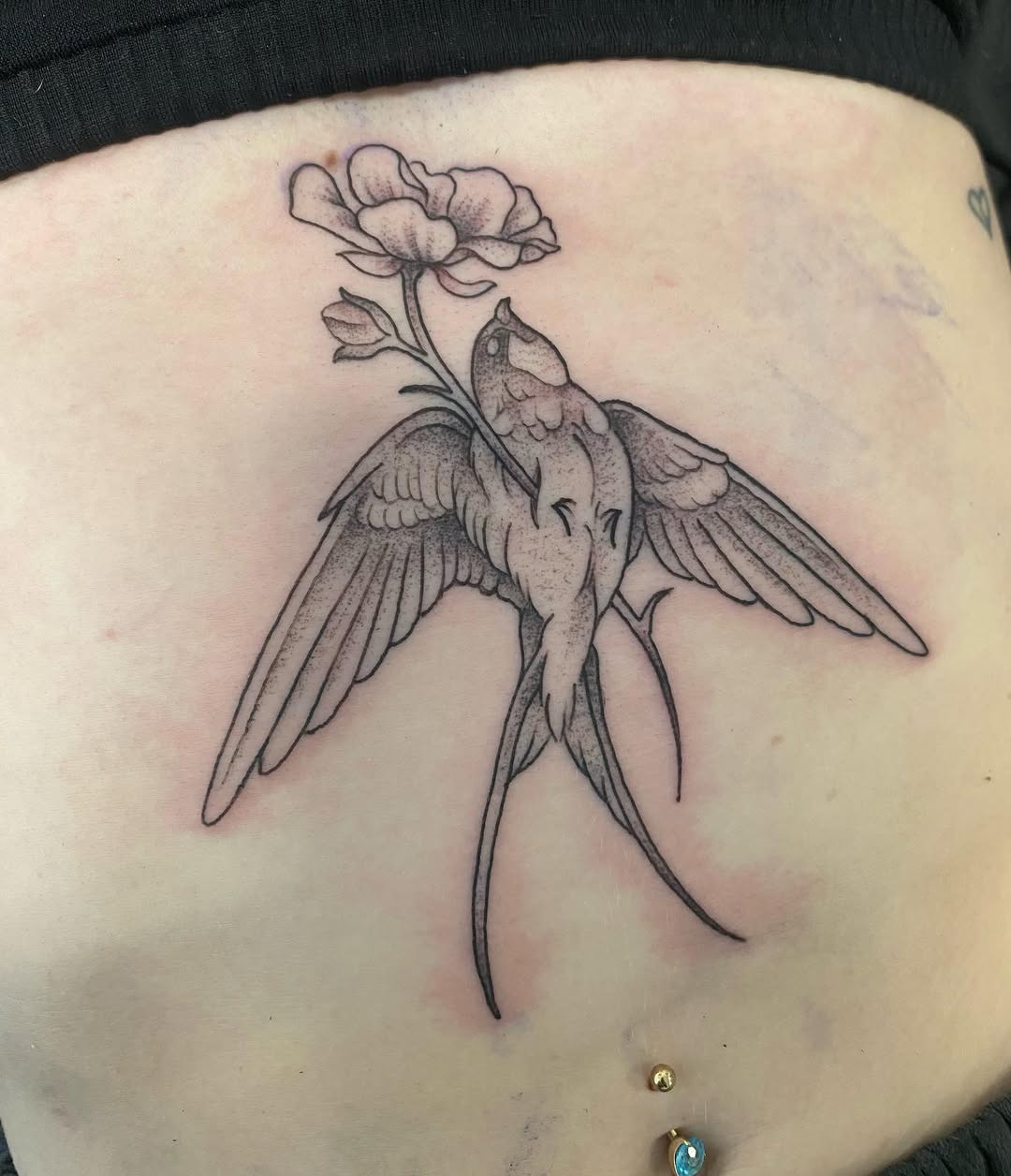 Winged bird with outstretched wings holding a delicate flower stem in its beak, rendered in black ink on upper back skin.