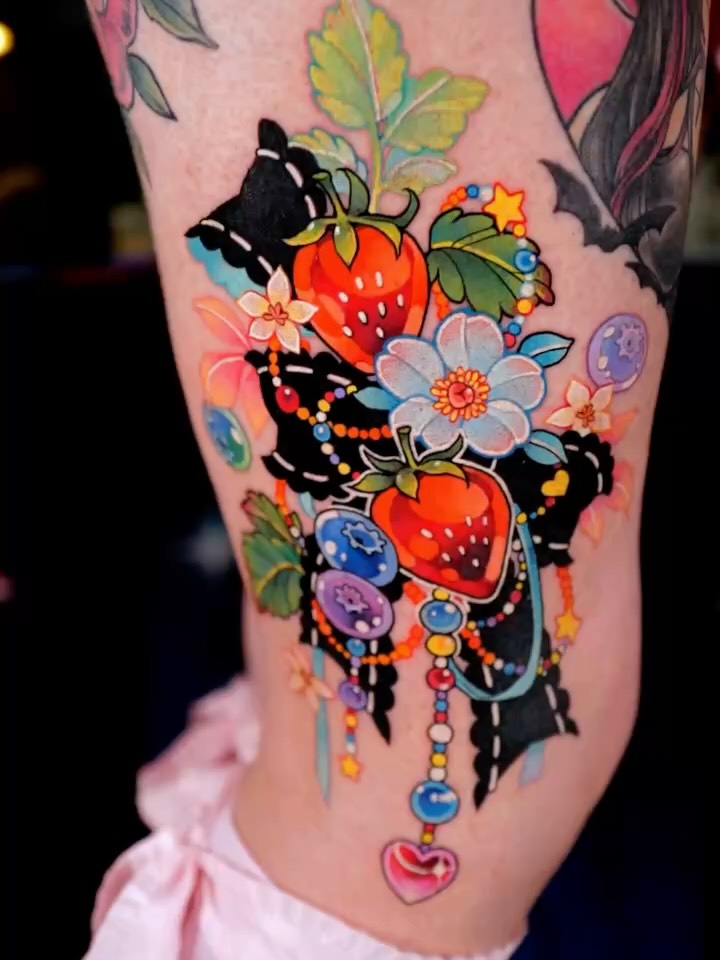 Colorful leg tattoo featuring strawberries, flowers, butterflies, and dangling beaded ornaments with black ribbon accents on pink skin.
