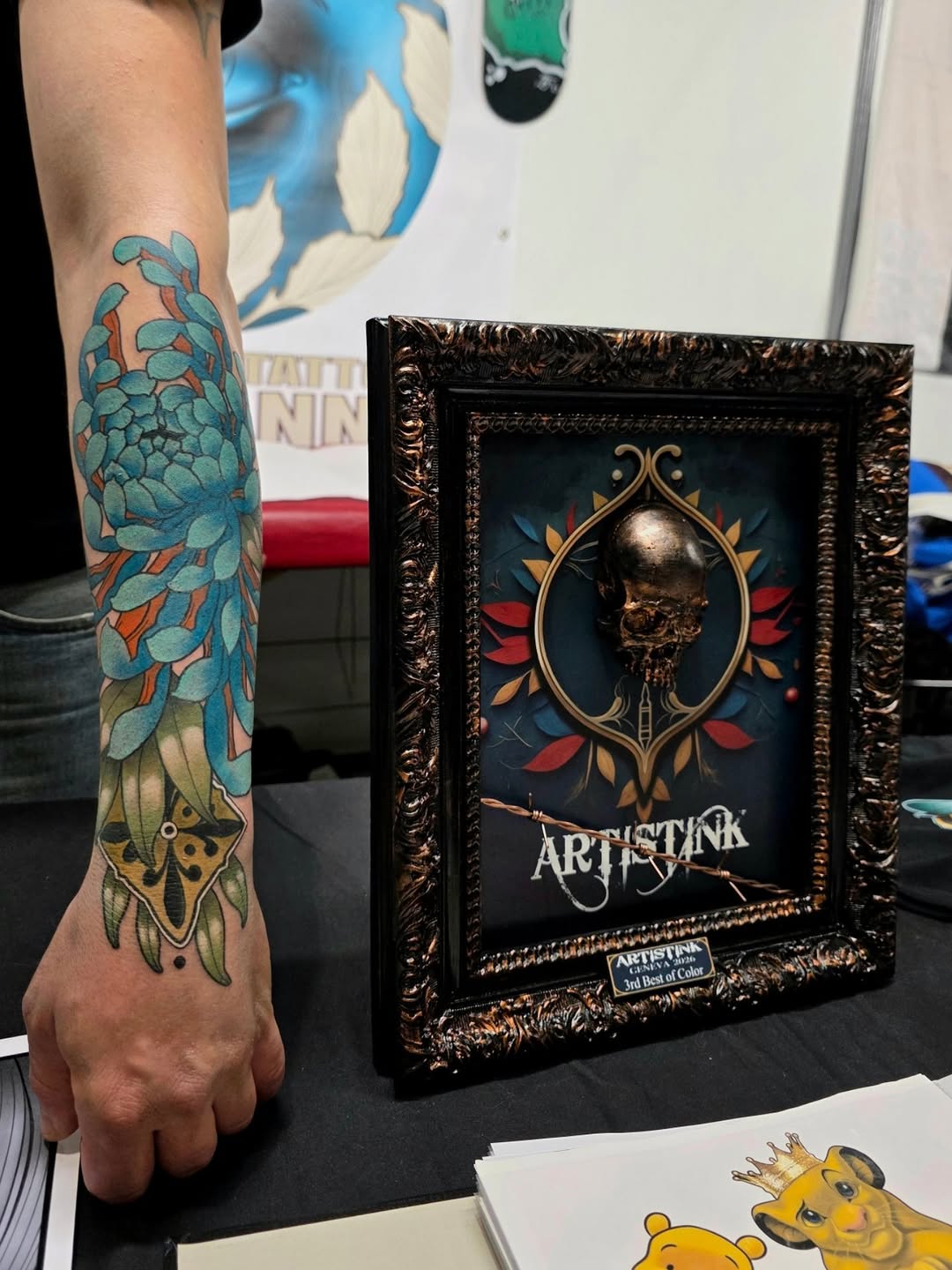 Colorful blue and yellow floral tattoo on forearm featuring layered petals and ornamental details, displayed next to a framed artwork.