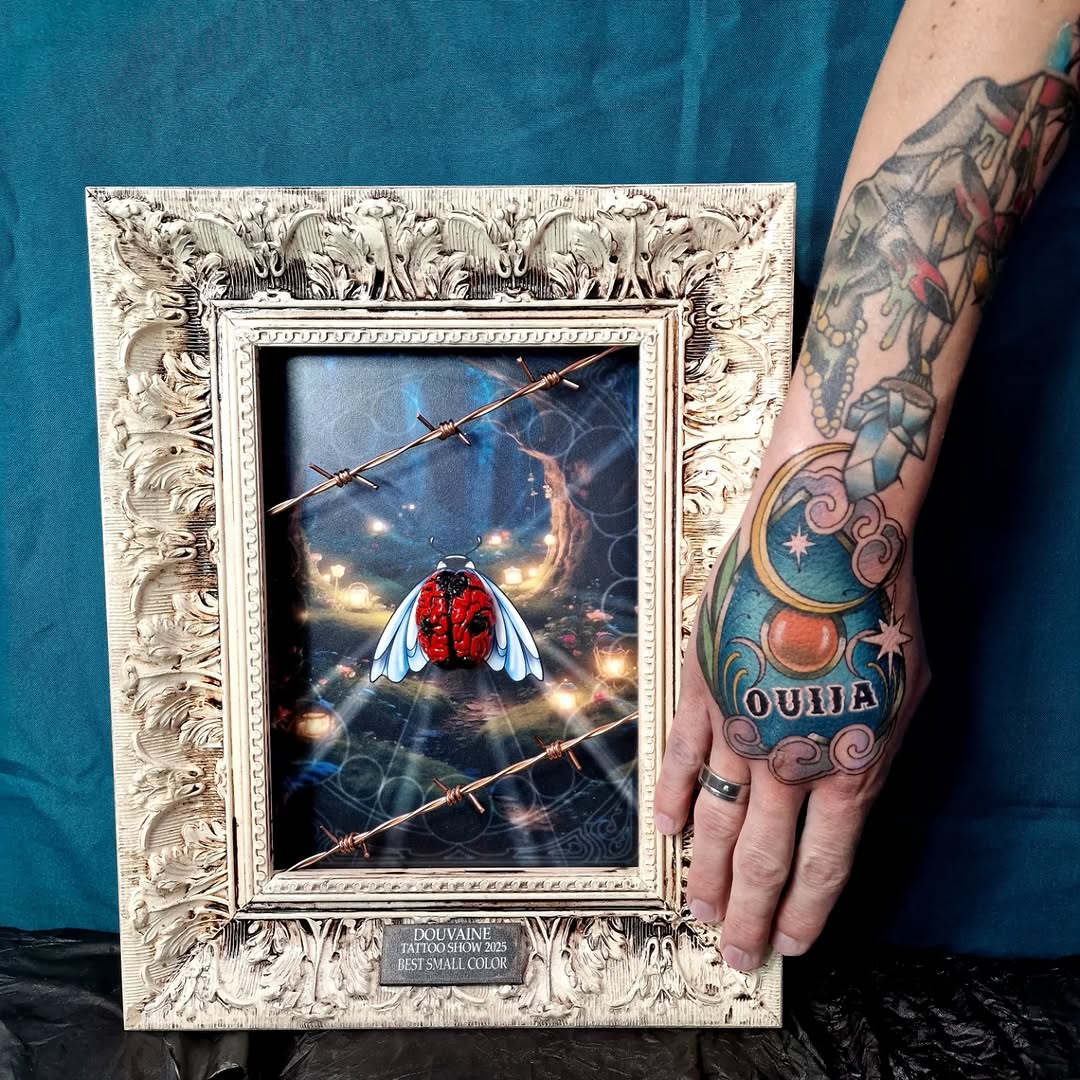Framed award photo displaying a hand tattoo featuring a red and white ladybug with barbed wire and a crescent moon design in blue, red, and gold colors.