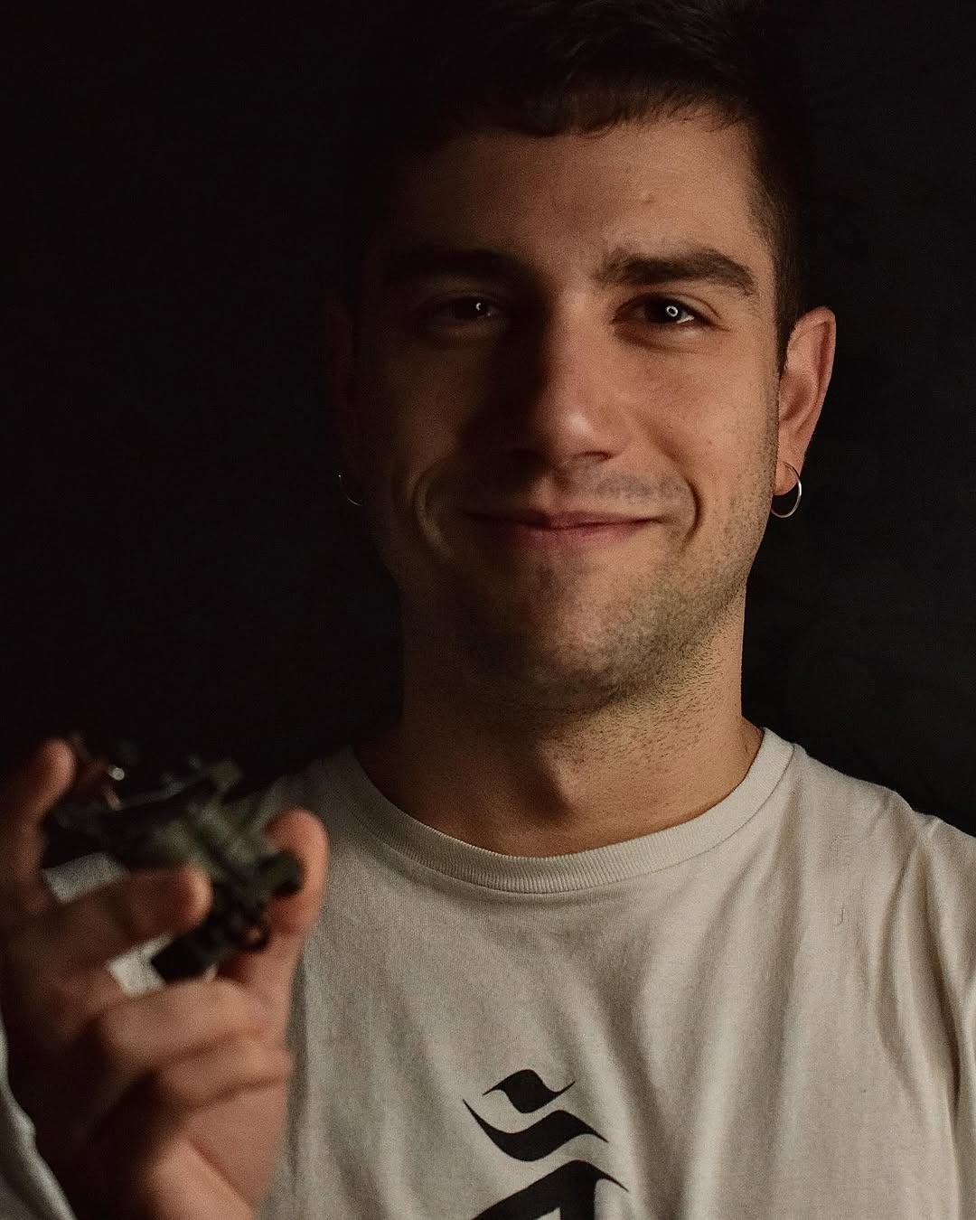 Man wearing cream t-shirt displaying a black wave logo tattoo on his chest, shown in portrait lighting against dark background.