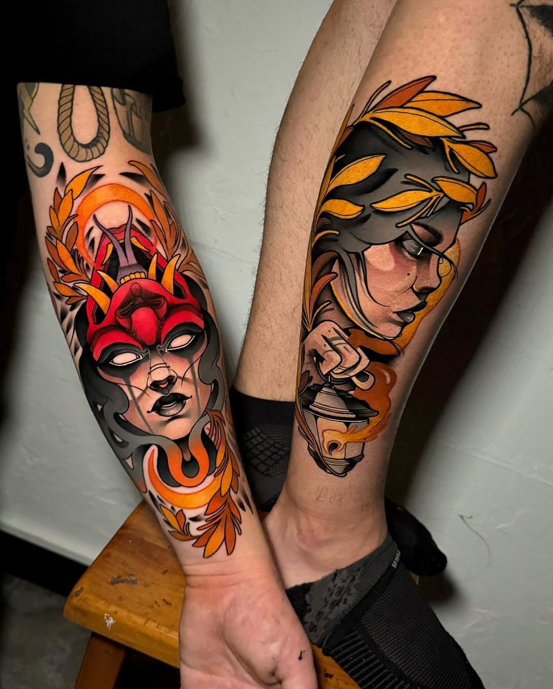 Two forearms display vibrant masked female faces with orange and red feathered headdresses, surrounded by decorative floral and serpent motifs in orange, red, and black ink.
