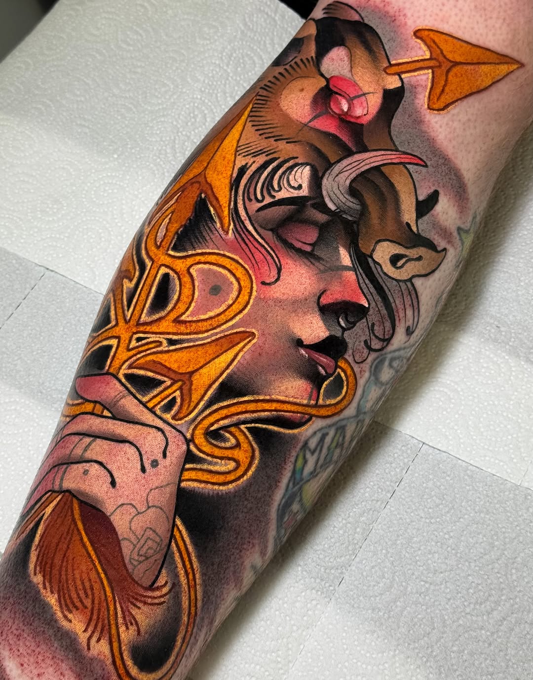Colorful tattoo on an arm featuring two female faces with ornate gold and orange decorative elements, pink shading, and flowing hair details.