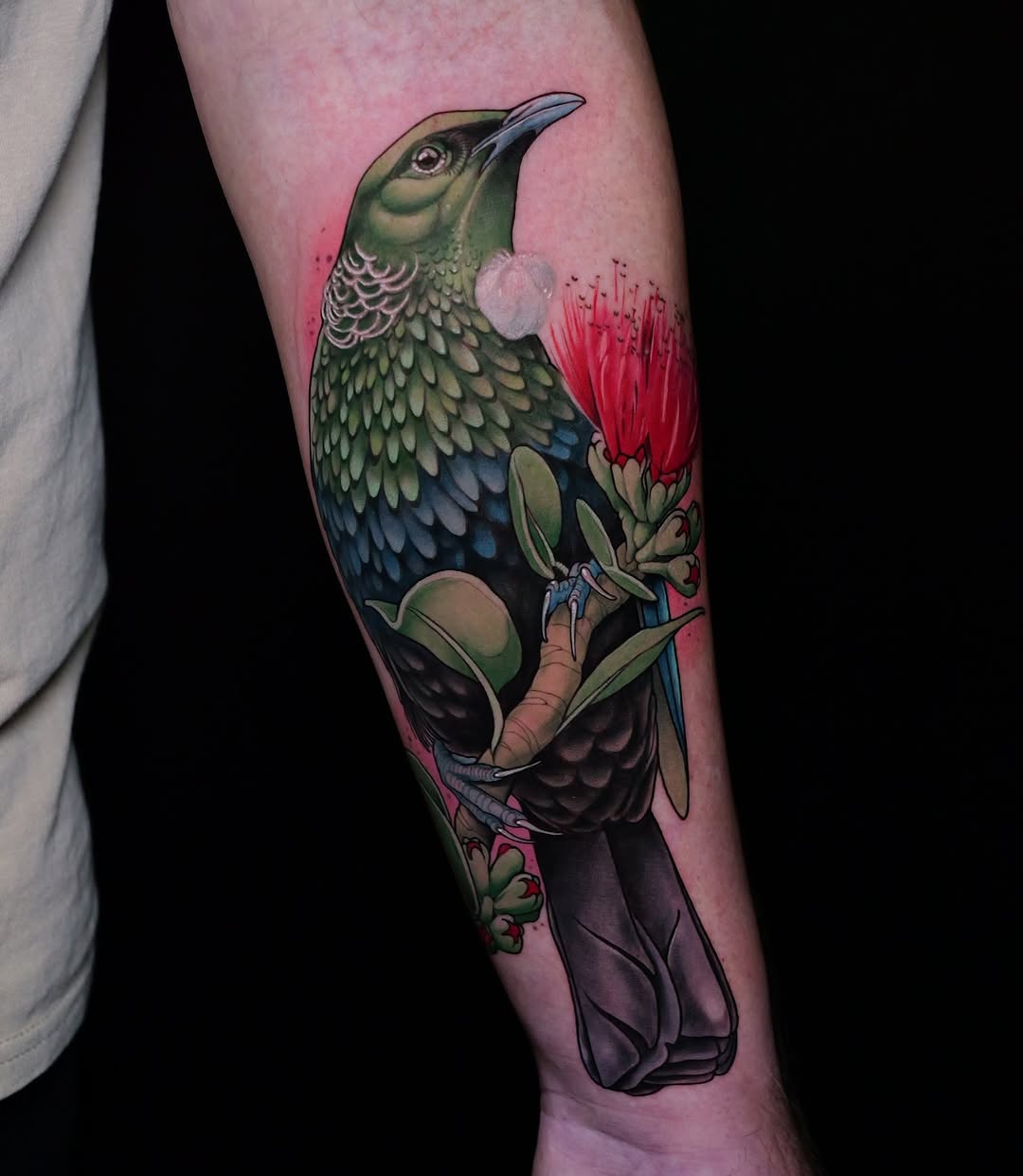 Colorful hummingbird tattoo holding a red flower on the forearm, featuring green, blue, and purple tones against pink skin.