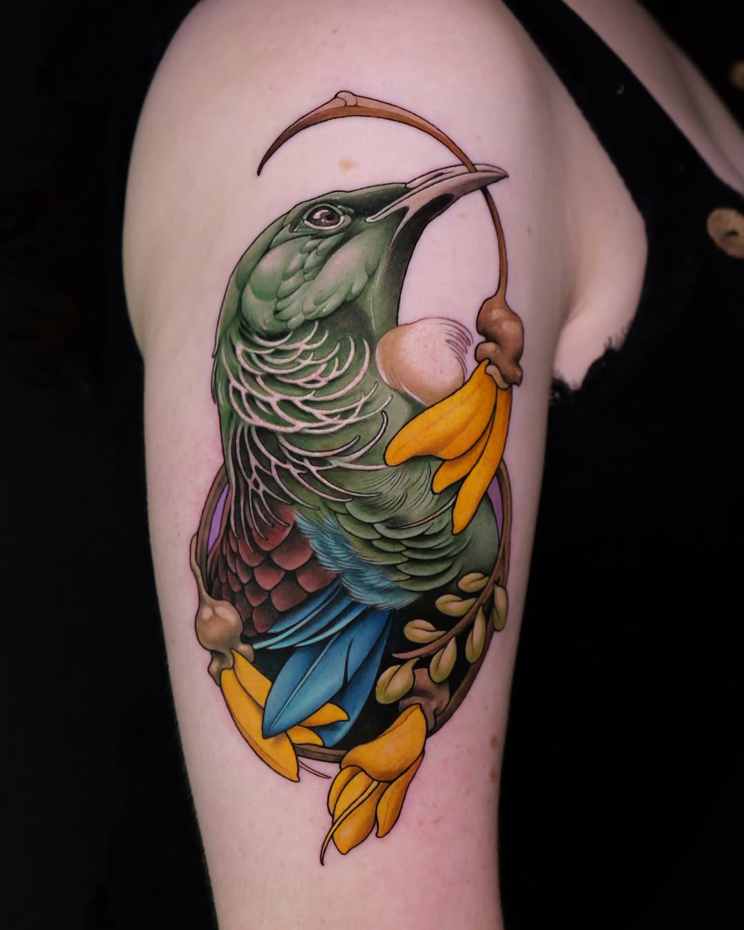 Colorful hummingbird tattoo on upper arm featuring green plumage, blue and red wing details, yellow feet, and a curved hook design above, on pale skin.