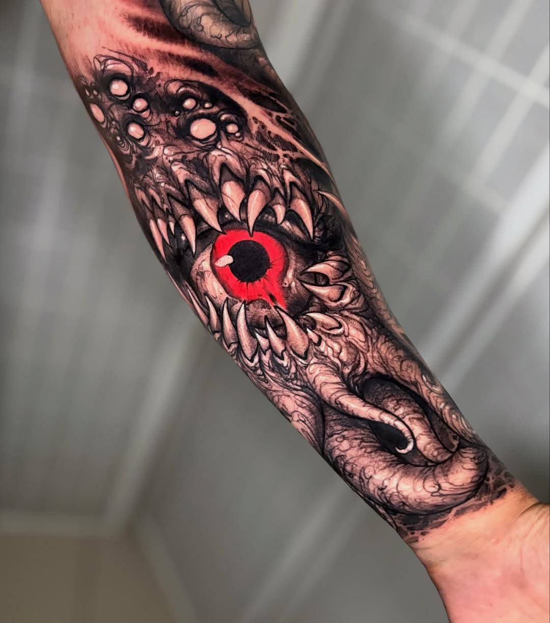 Detailed arm tattoo featuring a red-eyed creature with sharp teeth surrounded by ornamental patterns and organic shapes in black and red ink.