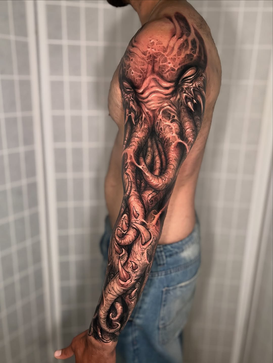Large-scale demon face and tentacle sleeve tattoo in red and black ink covering the shoulder, upper arm, and forearm of a shirtless man.