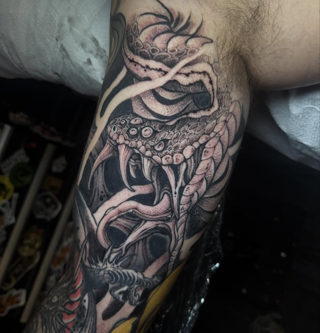 Detailed demon or oni mask tattoo on upper arm featuring intricate facial features, scales, and ornamental patterns in black and red ink.
