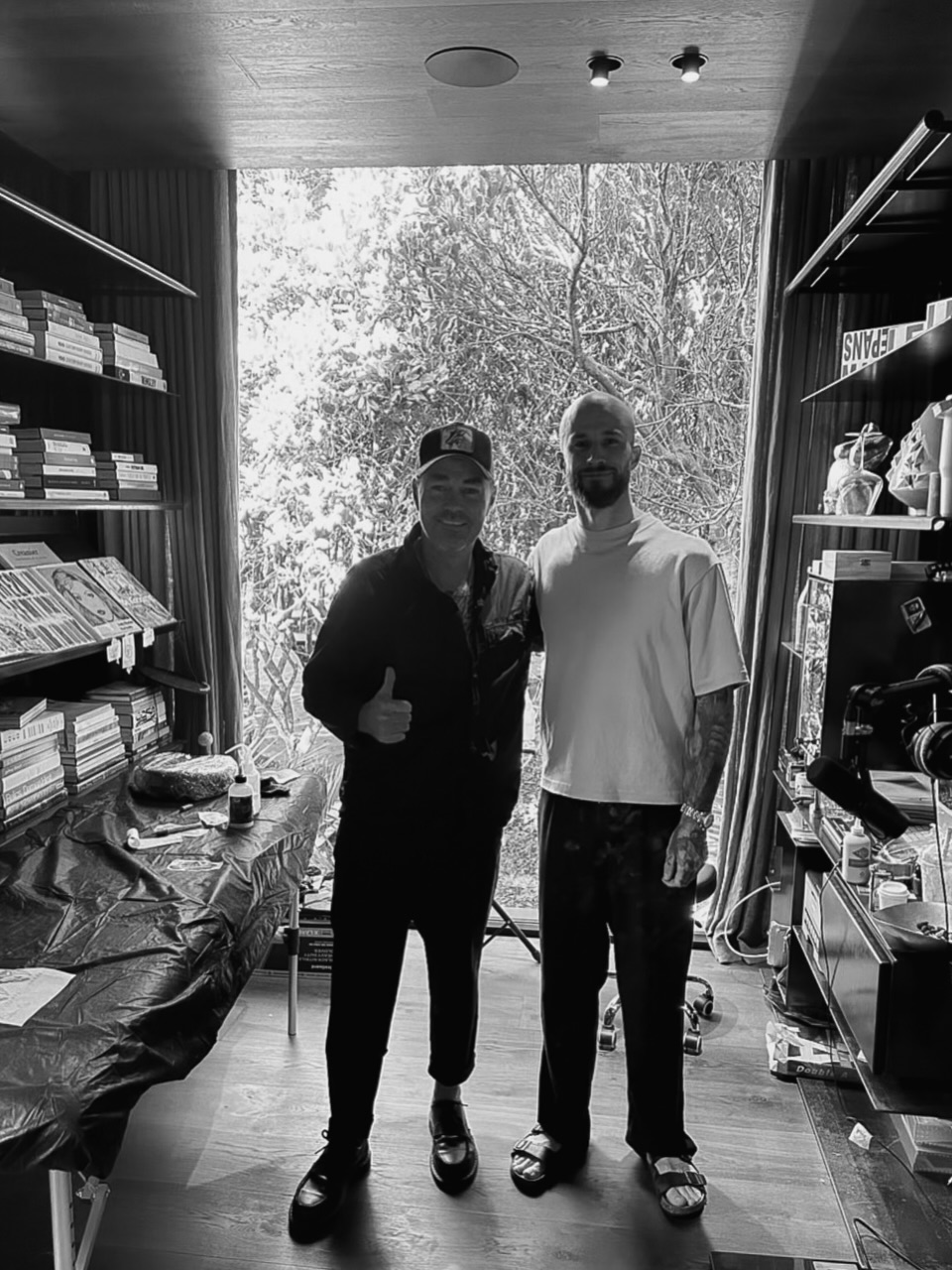 Two men stand in a retail shop with shelves and large windows showing trees; the man on the right displays an arm tattoo in dark ink.