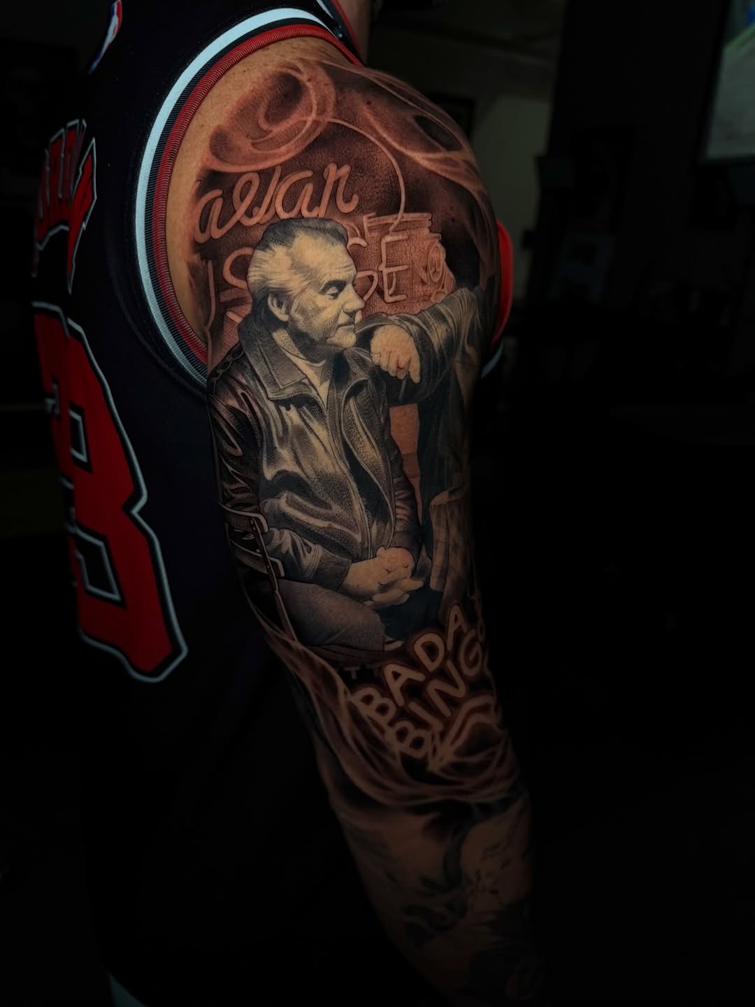 Portrait tattoo on upper arm showing a man in a jacket with his hand to his face, featuring red and brown tones with decorative text elements.