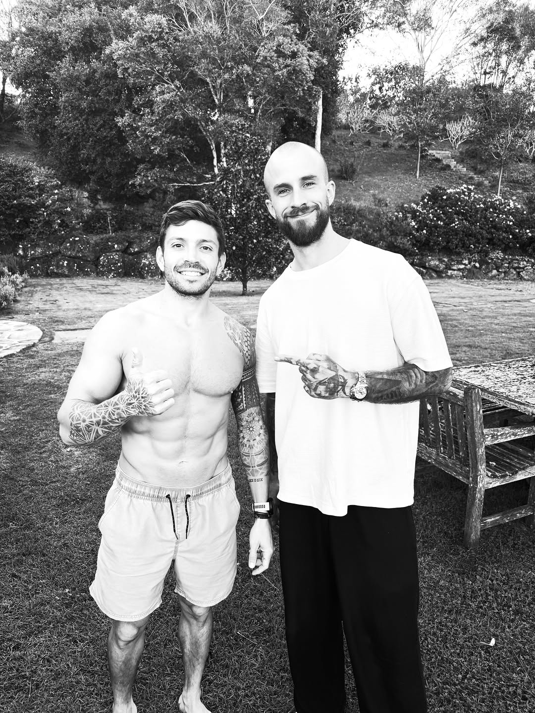 Two men posing outdoors, the man on the left displaying extensive tattoos on both arms and chest in a garden setting.