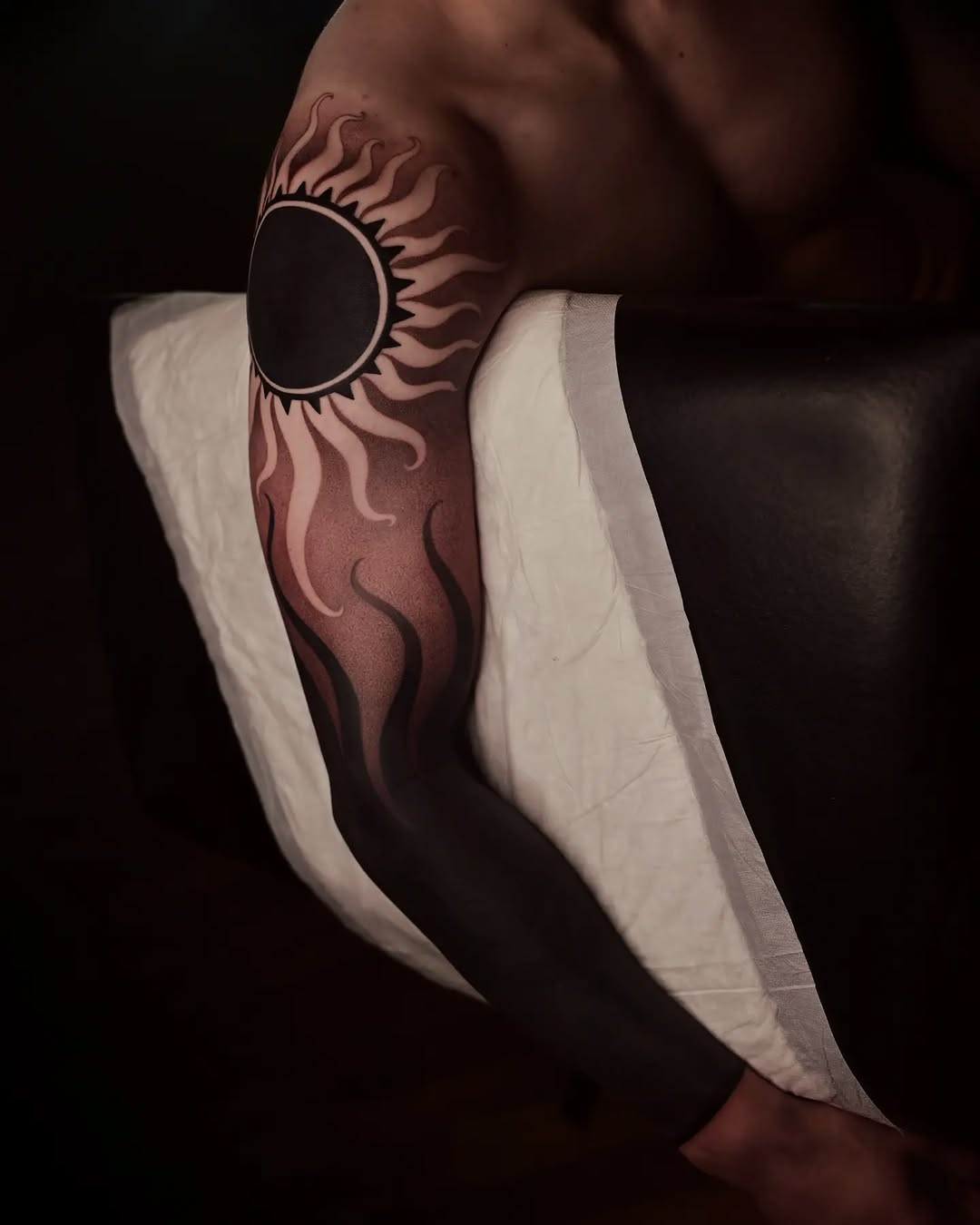Sun with radiating flame-like rays tattoo on upper arm in brown and black ink, displayed on skin with white gauze wrapping.