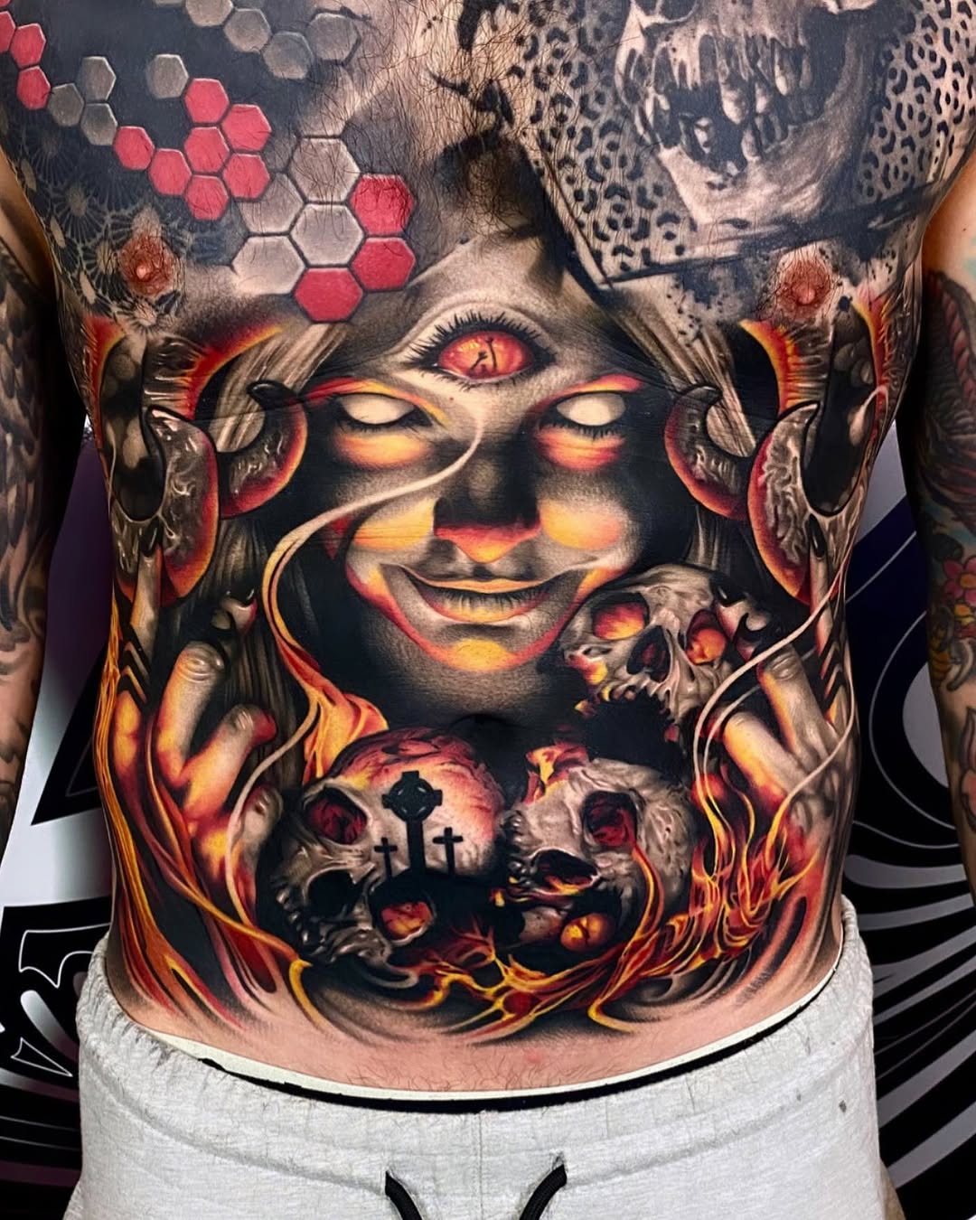 Colorful chest tattoo featuring a glowing-eyed face with flame accents, celestial patterns, skulls, and crosses in red, orange, and yellow tones.