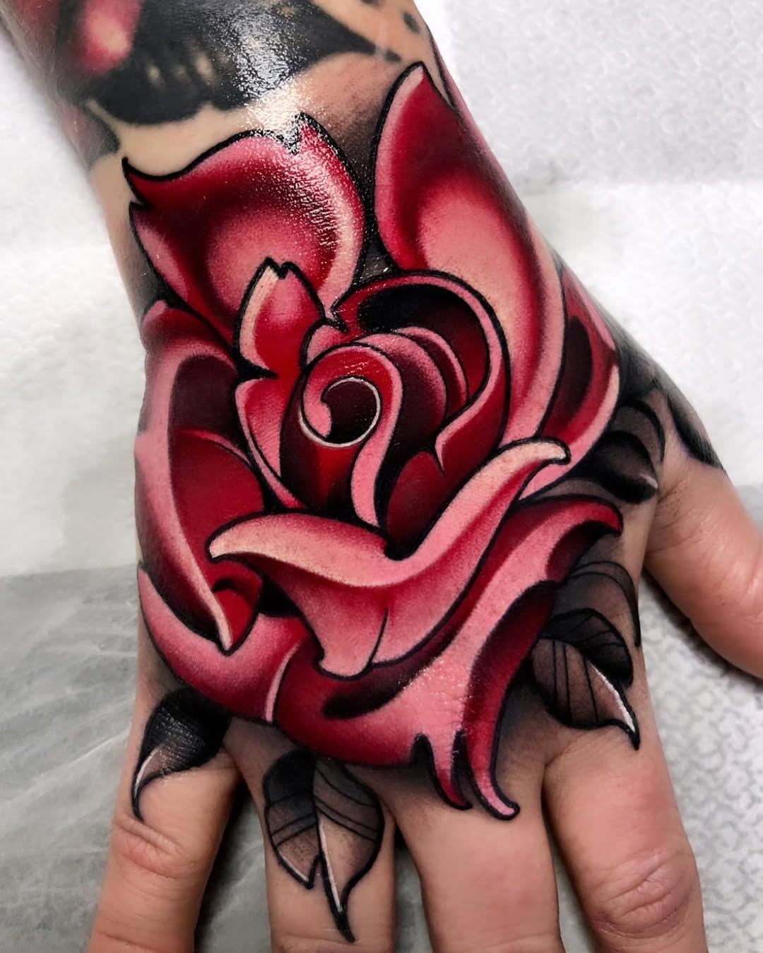 Red rose with black outline and shading on the back of a hand, featuring layered petals and green leaves.