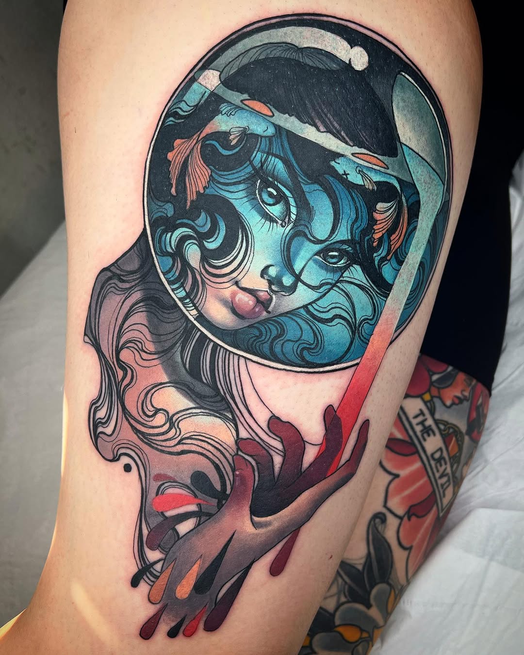 Colorful tattoo on upper arm featuring a figure with curled hair inside a circular frame, held by hands below in shades of blue, red, and pink.