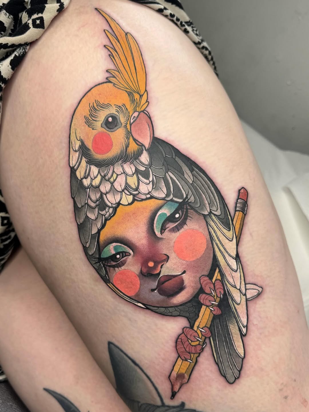 Colorful shoulder tattoo featuring a woman's face with red cheeks and teal eyeshadow, topped with a crested cockatiel perched on her head.