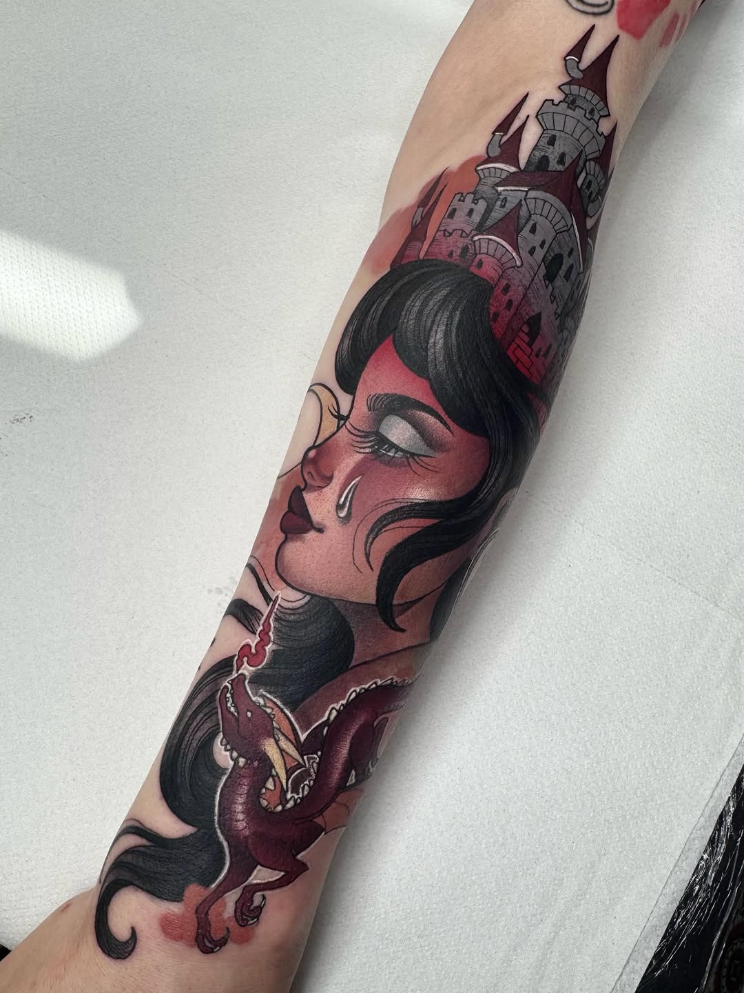 Color tattoo on forearm featuring a woman's profile with black hair, red and burgundy tones, a castle with spires, and a red demon figure below.